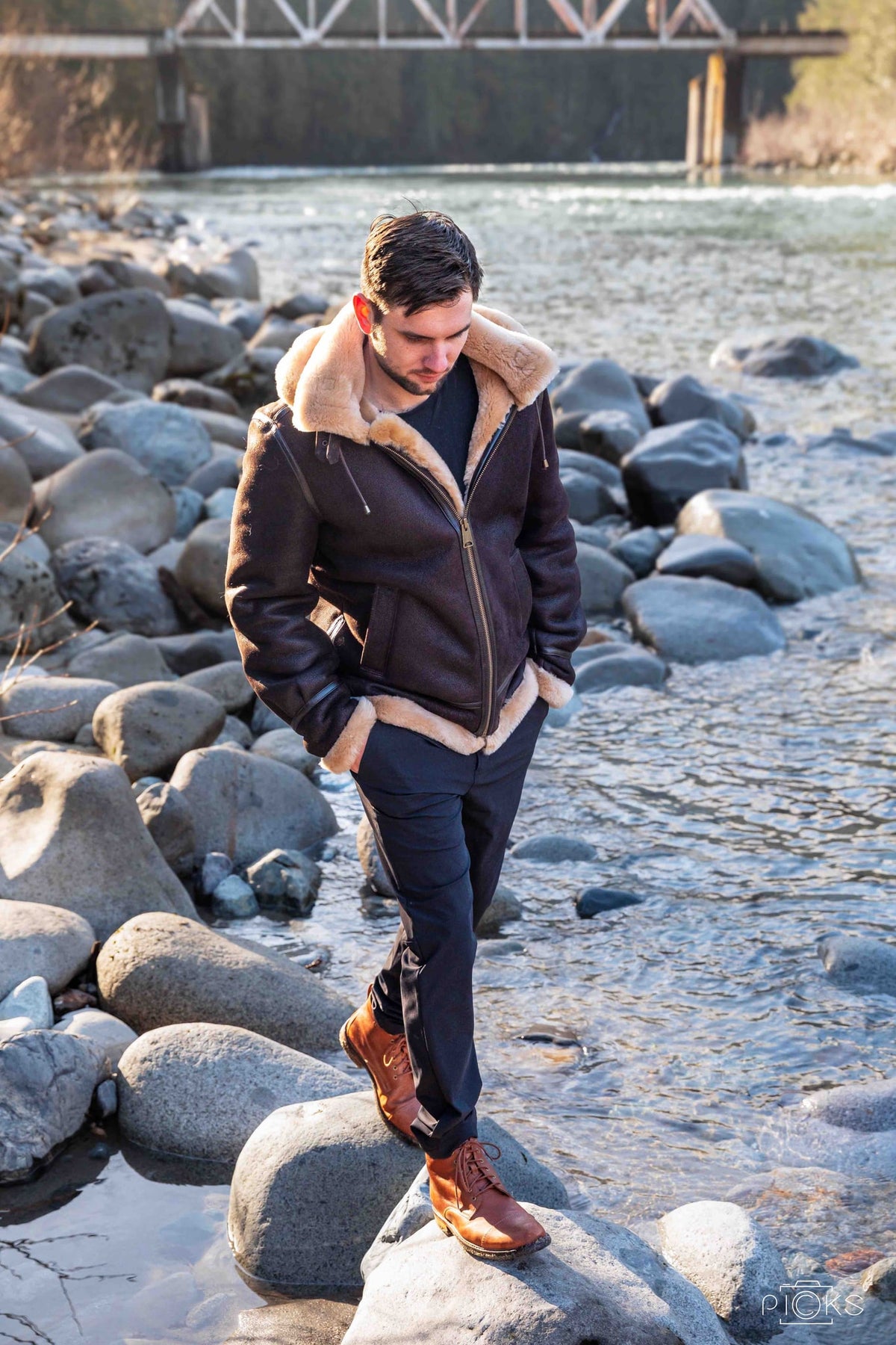Man in a sheepskin bomber jacket walking on a rocky riverbank