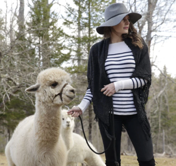 Woman in a striped shirt and wide-brimmed hat interacting with alpacas in a forested area