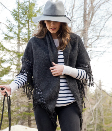 Woman wearing a dark gray fringe poncho, striped shirt, and hat outdoors with trees in the background